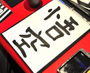 A guest at Shodocafe7557 in Roppongi, Tokyo, holds a calligraphy paper inscribed with the kanji ‘悟空’ (Goku), with an inkstone and brush on a red table.