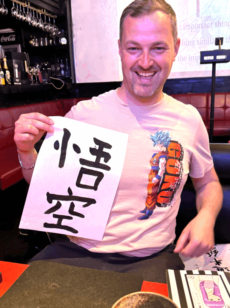 A solo guest at Shodocafe7557 in Roppongi, Tokyo, proudly holds a calligraphy paper displaying the kanji ‘悟空’ (Goku), with calligraphy tools and a projection screen in the background.