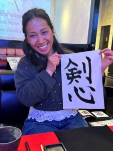 shodocafe7557 kakejiku single kenshin A solo guest at Shodocafe7557 in Roppongi, Tokyo, proudly displays a calligraphy sheet featuring the kanji ‘剣心’ (Kenshin), with calligraphy tools and a projection screen in the background.