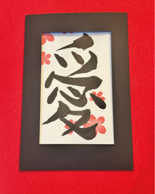 Calligraphy postcard featuring the kanji ‘ai’ (愛), decorated with cherry-blossom stamps in a black frame at Shodocafe7557 in Roppongi, Tokyo.