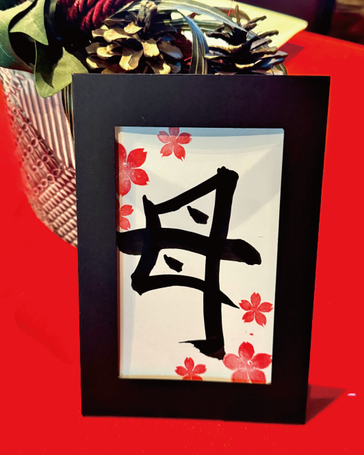 Calligraphy postcard featuring the kanji ‘haha’ (母), decorated with cherry-blossom stamps in a black frame at Shodocafe7557 in Roppongi, Tokyo.