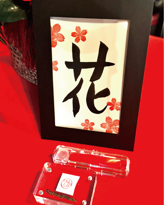 Calligraphy postcard featuring the kanji ‘hana’ (花), decorated with cherry-blossom stamps and bamboo accents in a black frame at Shodocafe7557 in Roppongi, Tokyo.