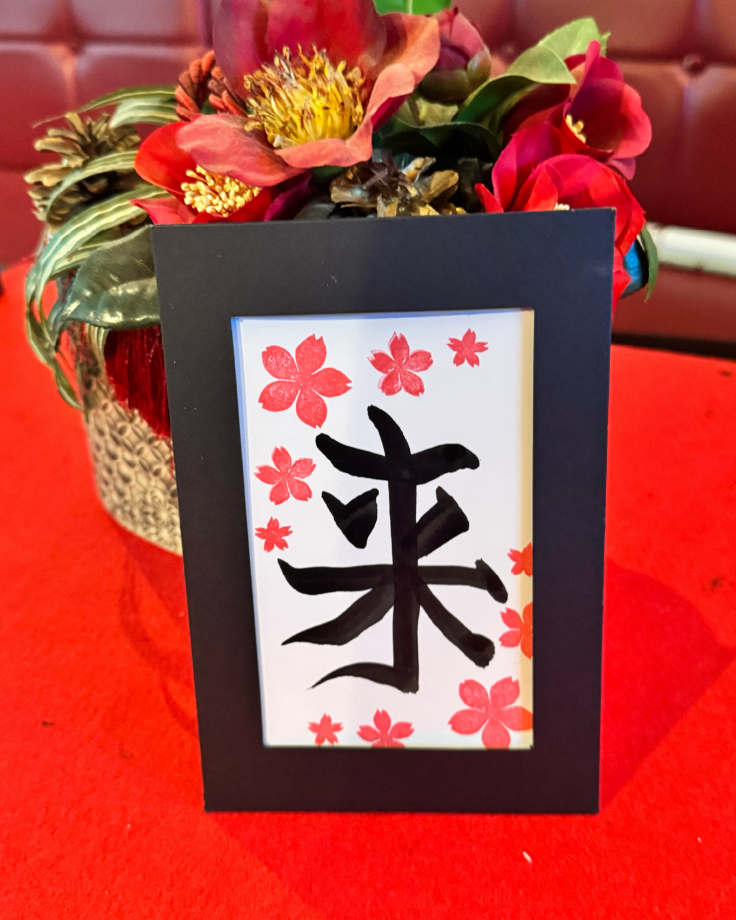 Calligraphy postcard featuring the kanji ‘rai’ (来), decorated with cherry-blossom stamps and bamboo accents in a black frame at Shodocafe7557 in Roppongi, Tokyo.
