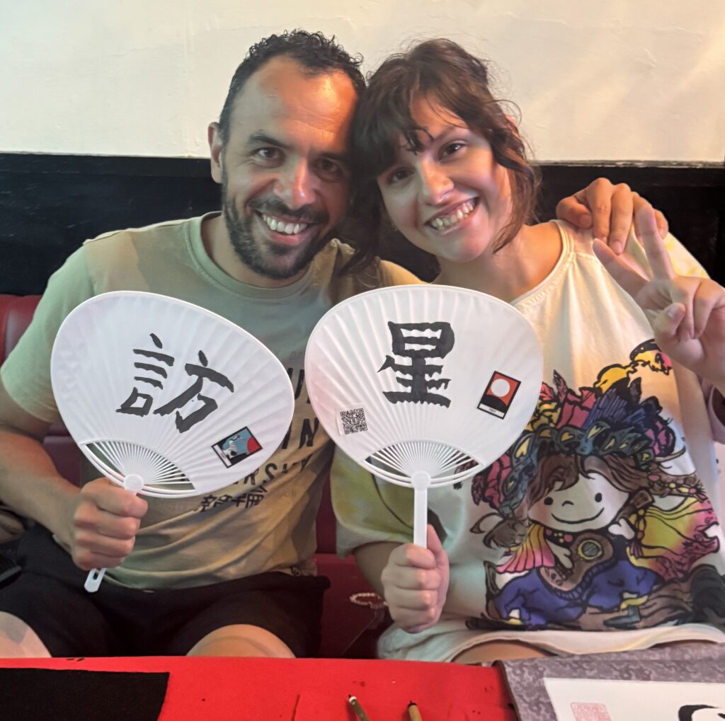 hotel calligraphy uchiwa visiting star A smiling couple at Shodocafe7557 in Tokyo holds hand-painted uchiwa fans featuring the kanji ‘訪’ (visit) and ‘星’ (star) created during the hotel-guest calligraphy workshop.