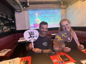 Newlyweds Celeste and David during their honeymoon calligraphy experience at Shodocafe7557 in Roppongi, Tokyo, smiling with uchiwa fans painted with kanji characters ‘岩’ (rock) and ‘友心’ (friendship & heart).