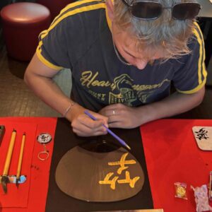 Hotel guest painting golden kanji characters on a traditional black uchiwa fan during the exclusive calligraphy workshop at Shodocafe7557 in Roppongi, Tokyo.