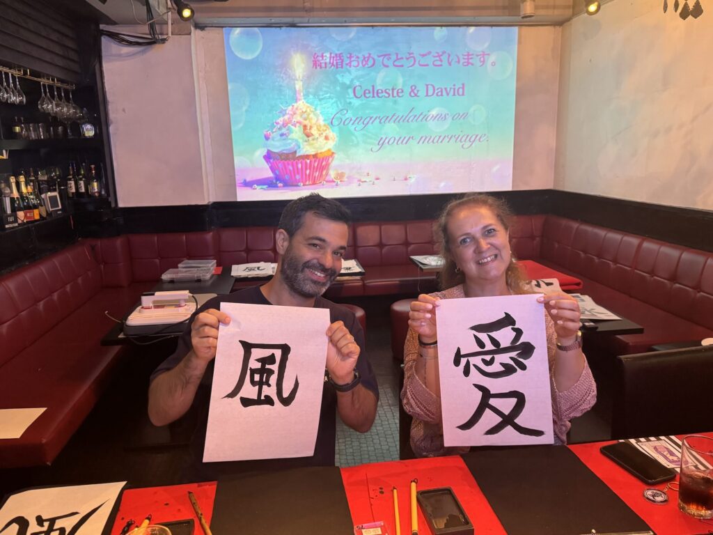 shodocafe7557 roppongi calligraphy newlyweds celeste david scroll Celeste and David painting kanji characters ‘愛 (Love)’ and ‘風 (Wind)’ on traditional uchiwa fans during their honeymoon calligraphy experience at Shodocafe7557.