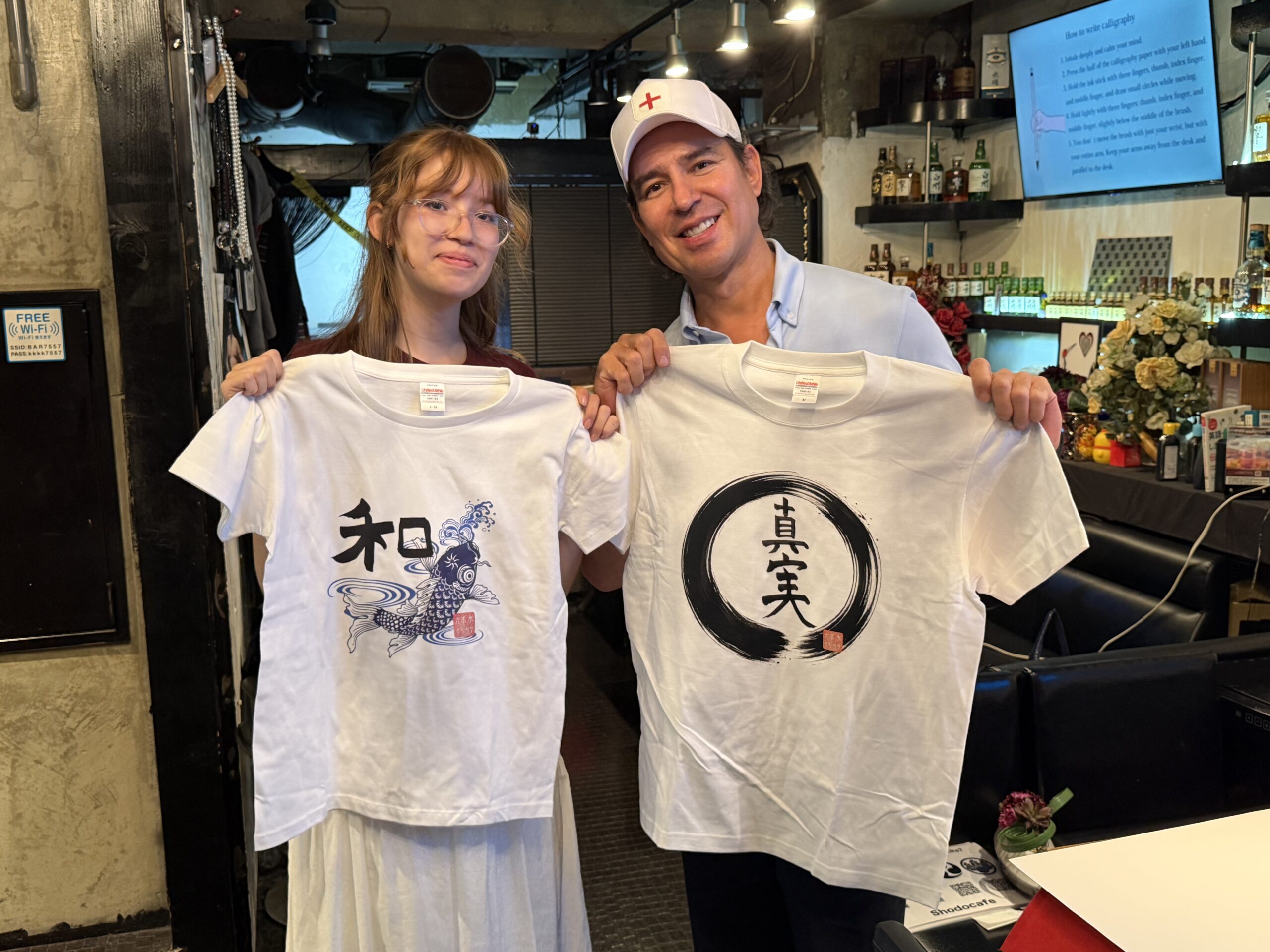 Guests holding their original handwritten kanji T-shirts after a Japanese calligraphy T-shirt experience at Shodocafe7557 in Roppongi, Tokyo